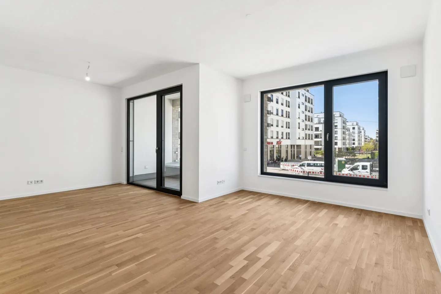 Bright, empty room with light wood floors, white walls, and black-framed windows and sliding glass door. Cityscape visible through the window.