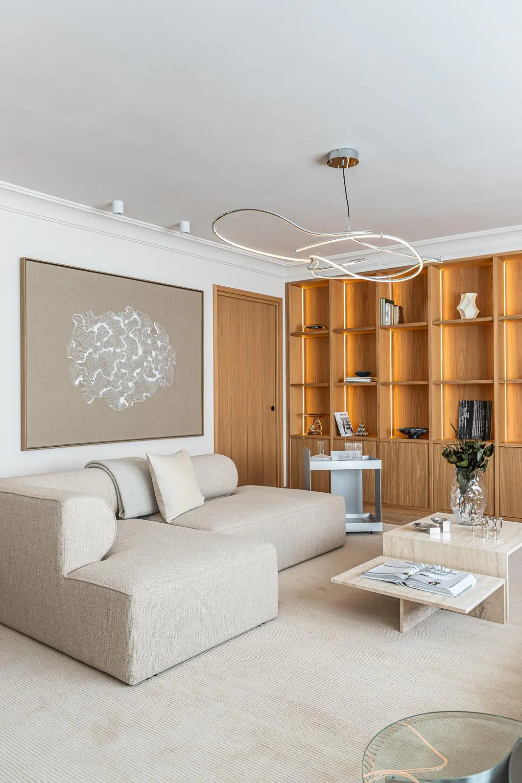 Bright living room with a beige sectional sofa, a tan painting, and a wooden bookshelf with warm lighting.