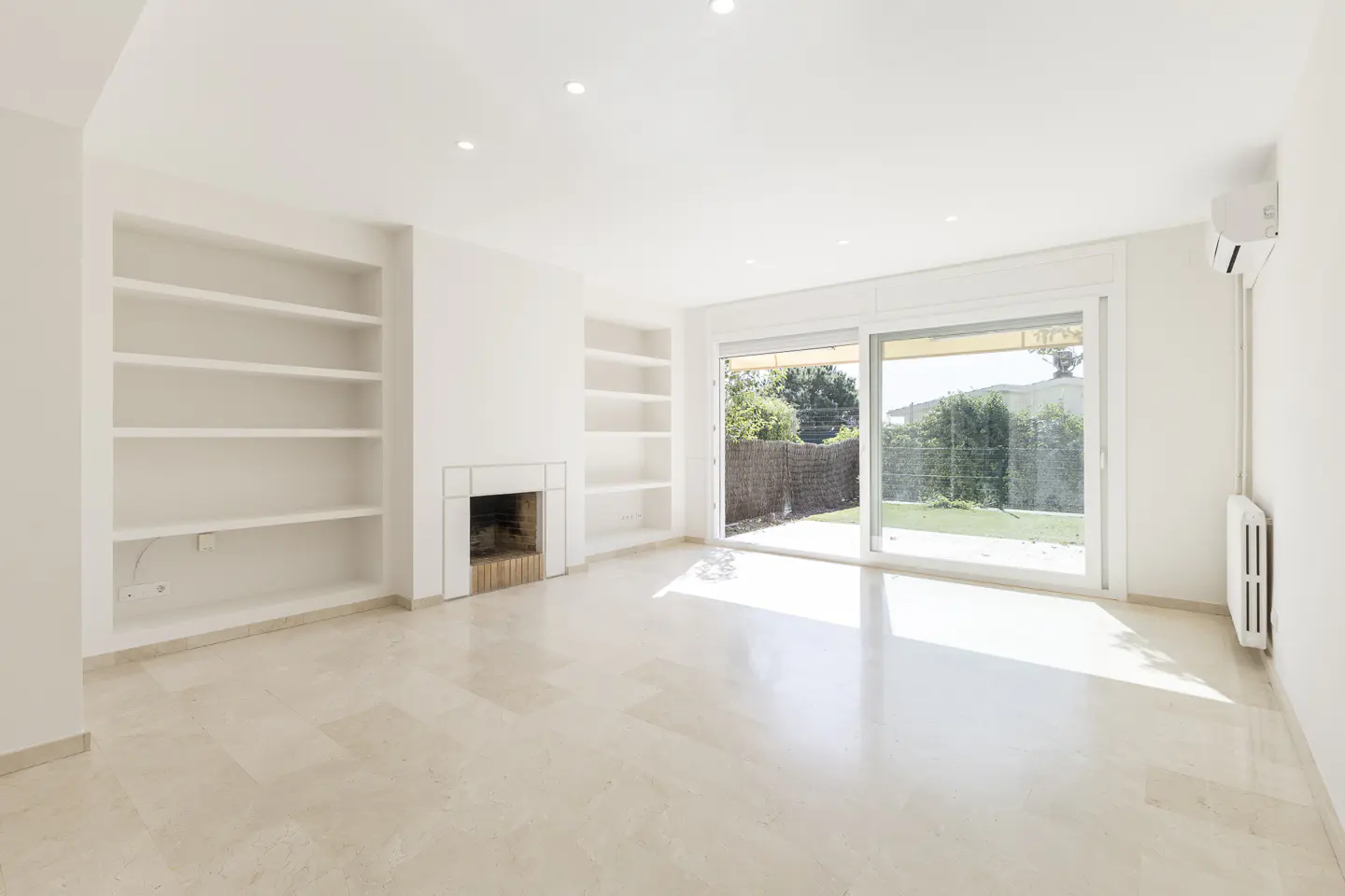 Bright, empty living room with marble floors, white walls, built-in shelves, fireplace, and sliding glass doors to a green backyard.