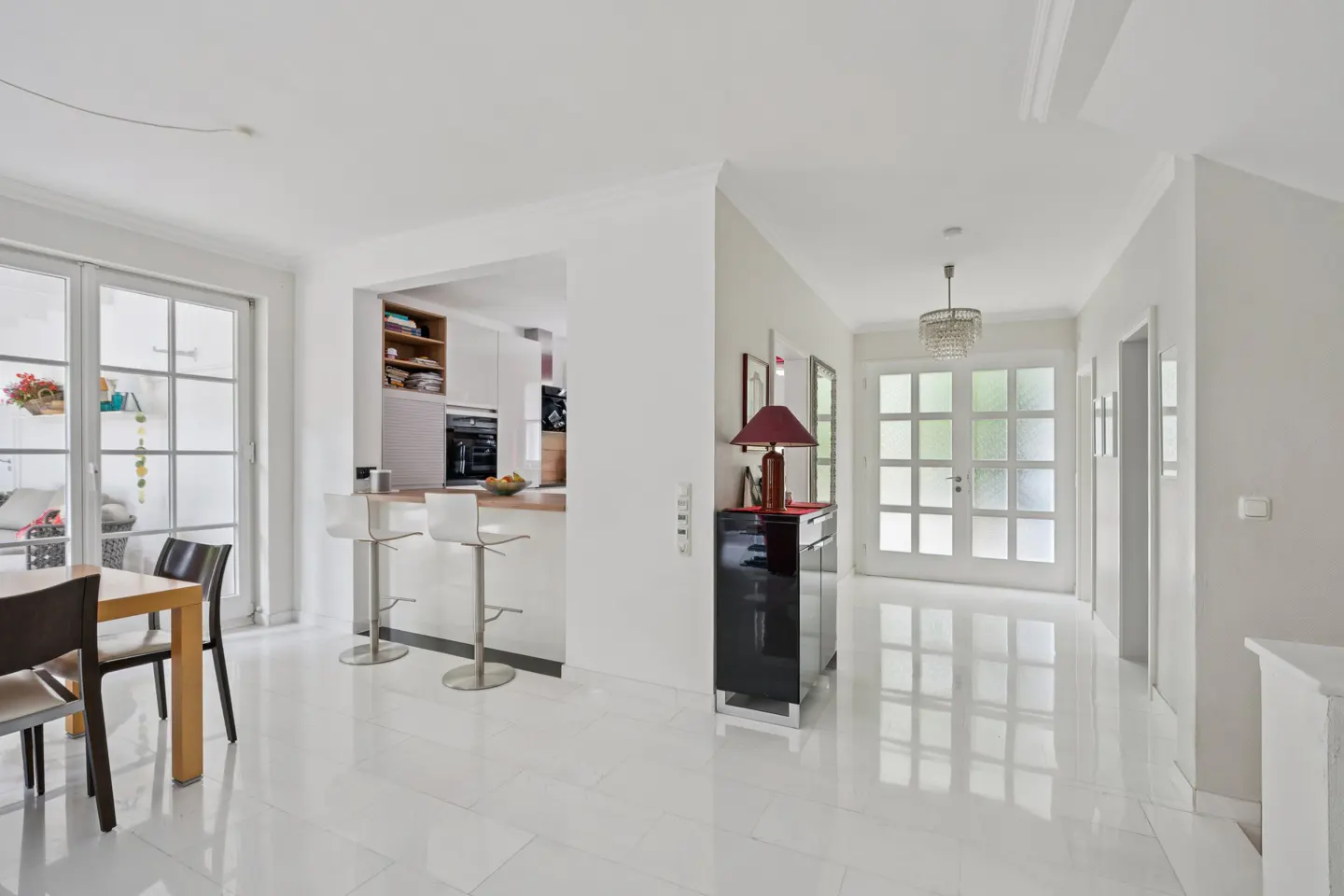 Bright, modern home interior with white tile floors, a kitchen with bar stools, and a dining table with chairs. A black cabinet and glass-paneled doors are visible.