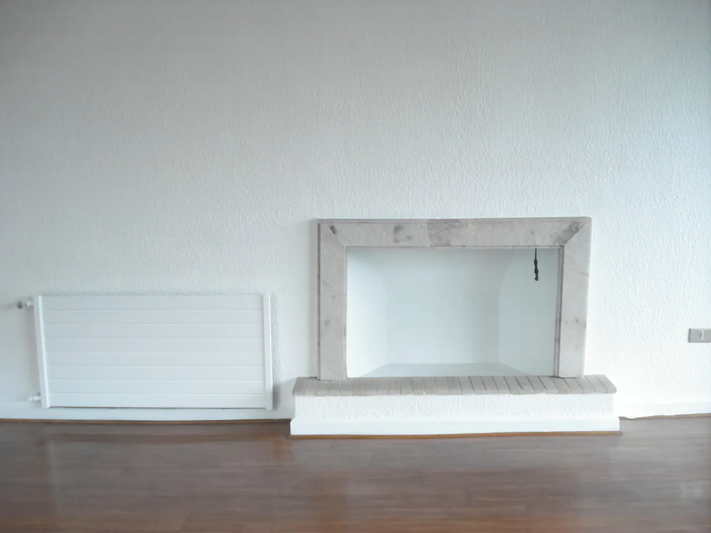 Empty room with wood floor, white walls, radiator, and a stone fireplace.