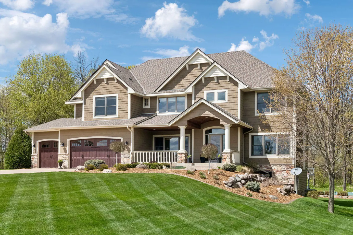 Two-story tan house with a brown roof, a front porch, and a green lawn on a sunny day.
