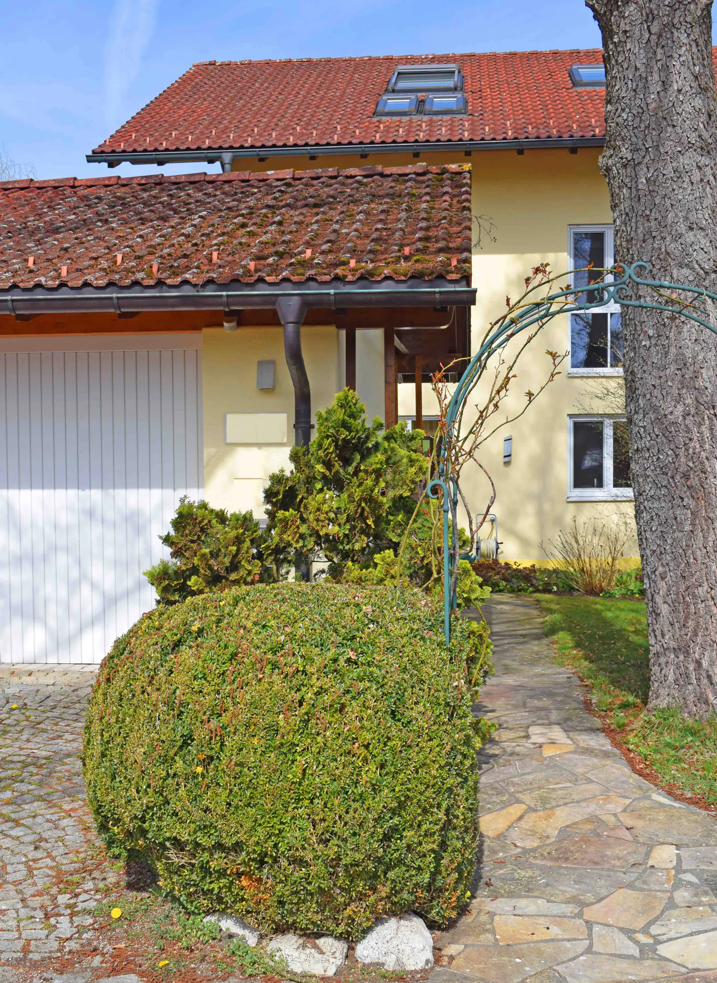 A yellow house with a red tile roof, a white garage door, and a stone walkway. Green bushes are in the front.