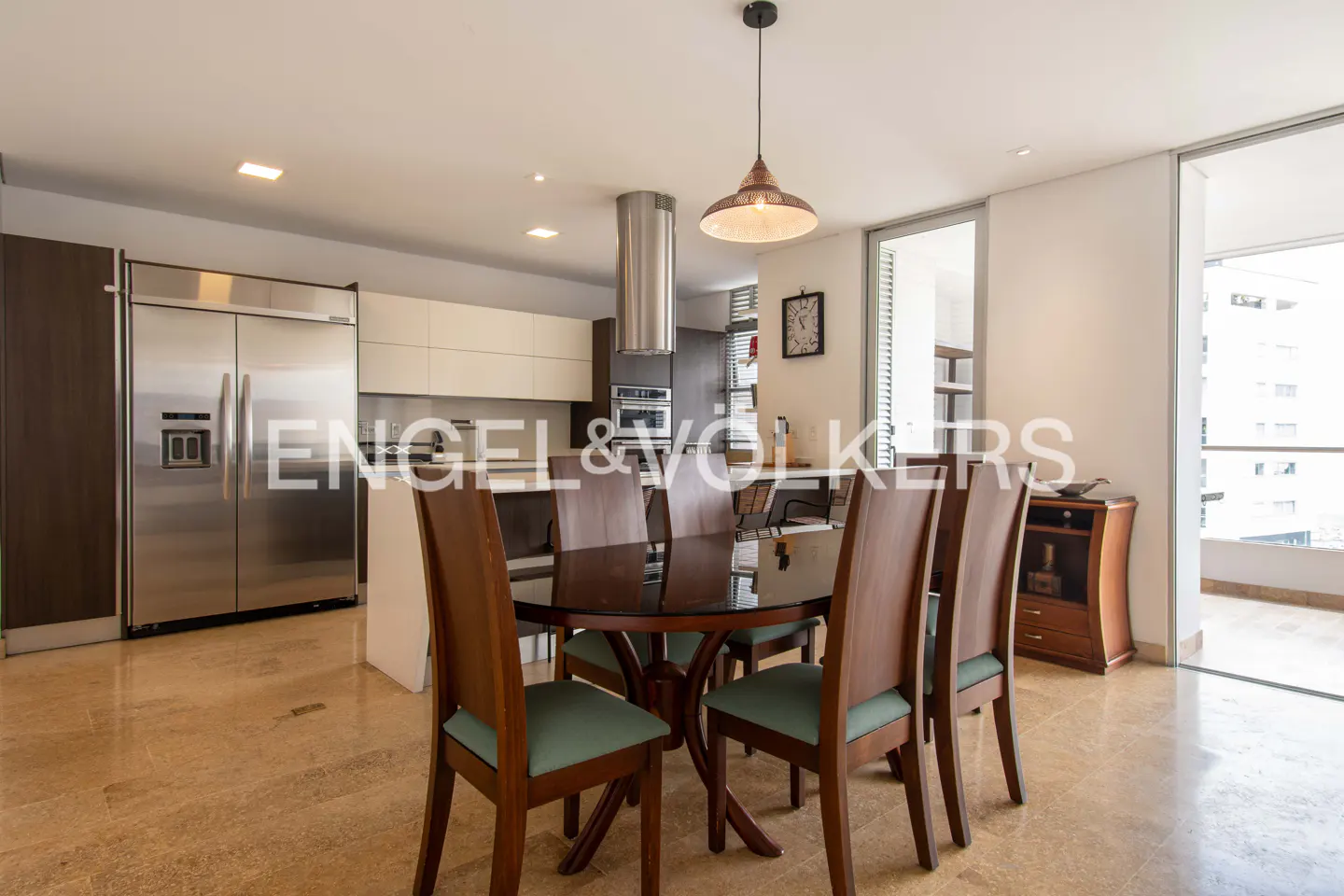 Modern dining area with a glass table, six wooden chairs with teal cushions, and an open kitchen with stainless appliances.