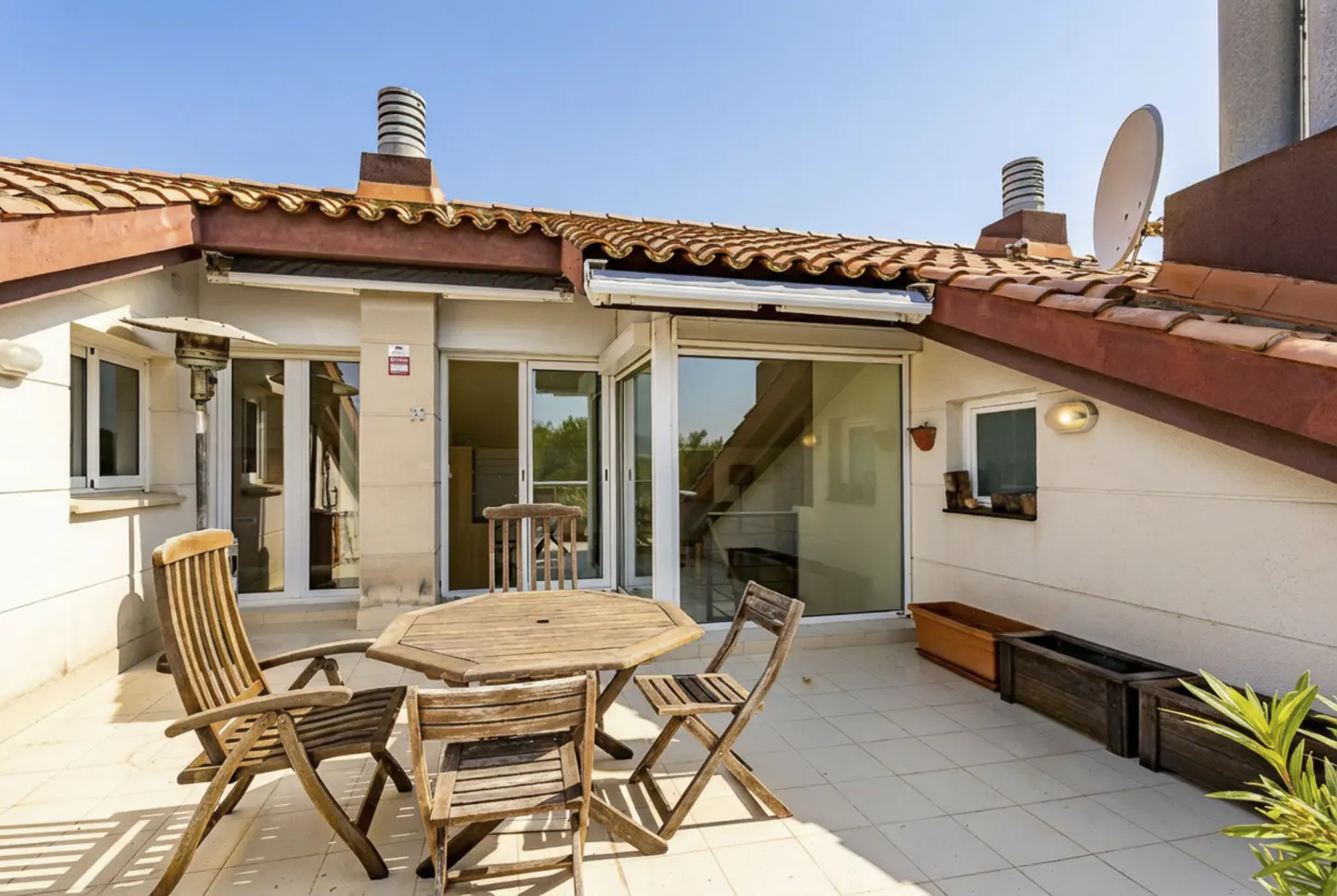 Outdoor patio with wooden table and chairs on a tiled floor, with a red-tiled roof and blue sky above.