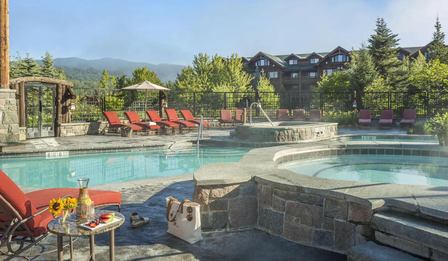 Outdoor pool area with red lounge chairs, a stone hot tub, and a table with drinks and snacks. A large lodge is in the background.