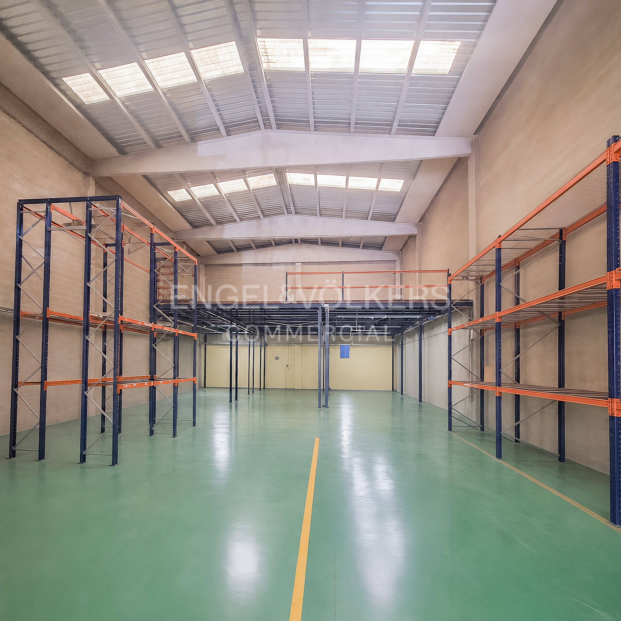 Empty warehouse interior with high ceilings, skylights, green epoxy floors, and tall blue and orange metal storage racks.