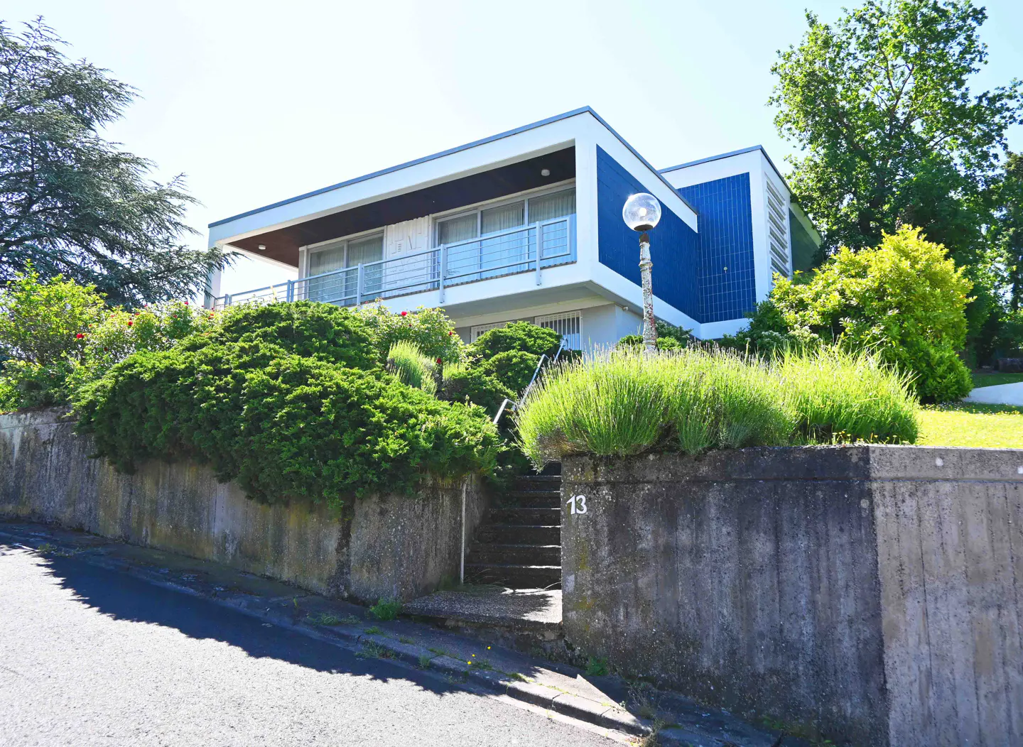 Modern two-story house with blue and white exterior, set on a hill with stone retaining wall and stairs leading up to the entrance.