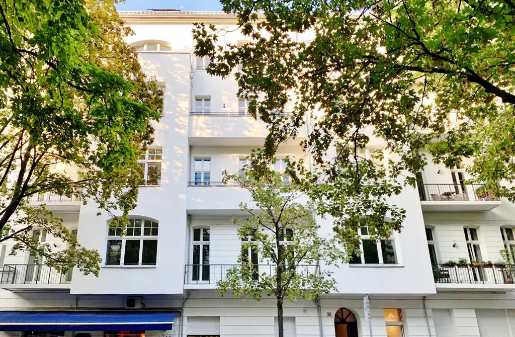 Exterior view of a white, multi-story apartment building with balconies, framed by green trees.