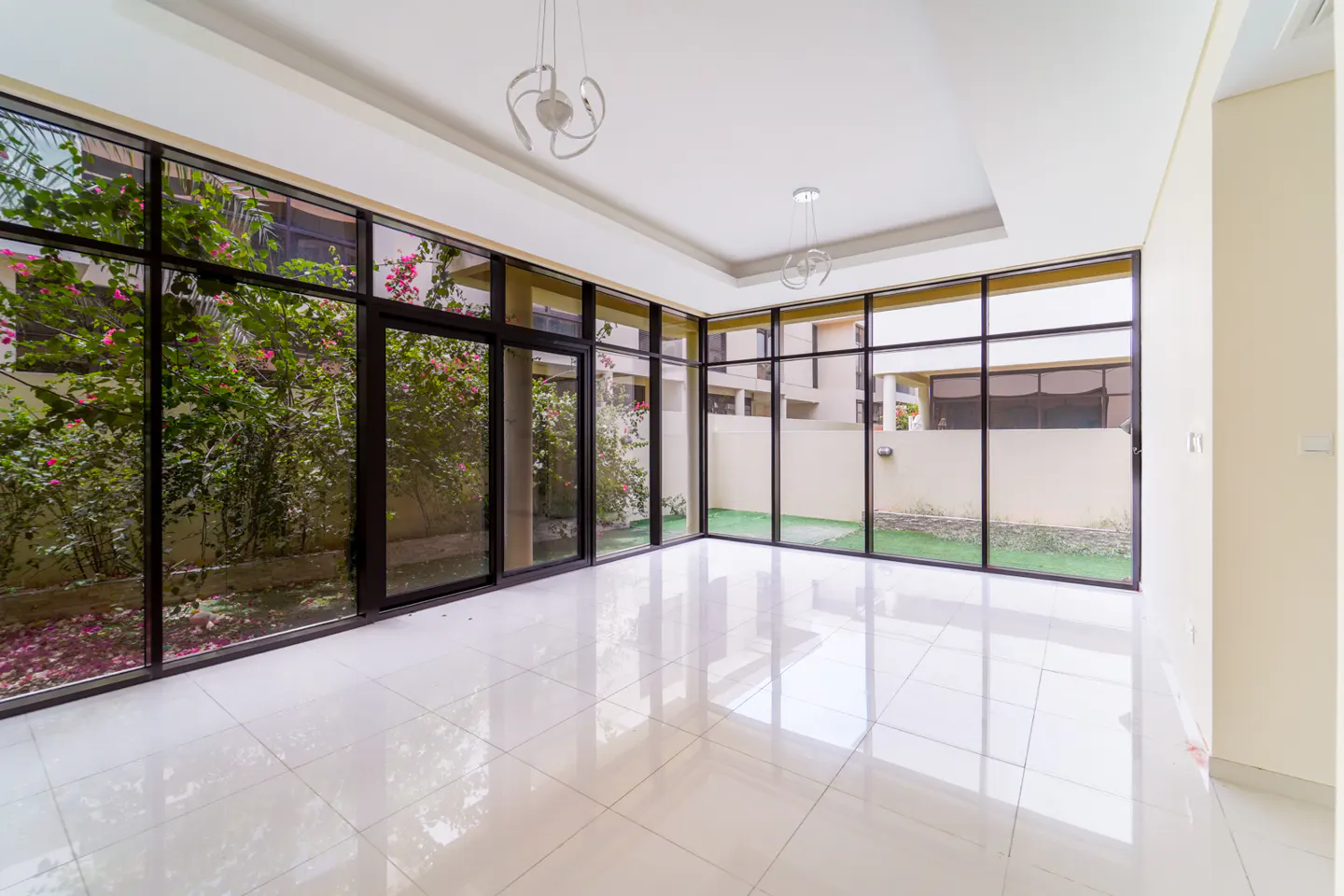 Bright, empty room with white tile floor and black-framed windows. Outside, lush greenery and a glimpse of neighboring buildings. Modern chandeliers hang from the ceiling.