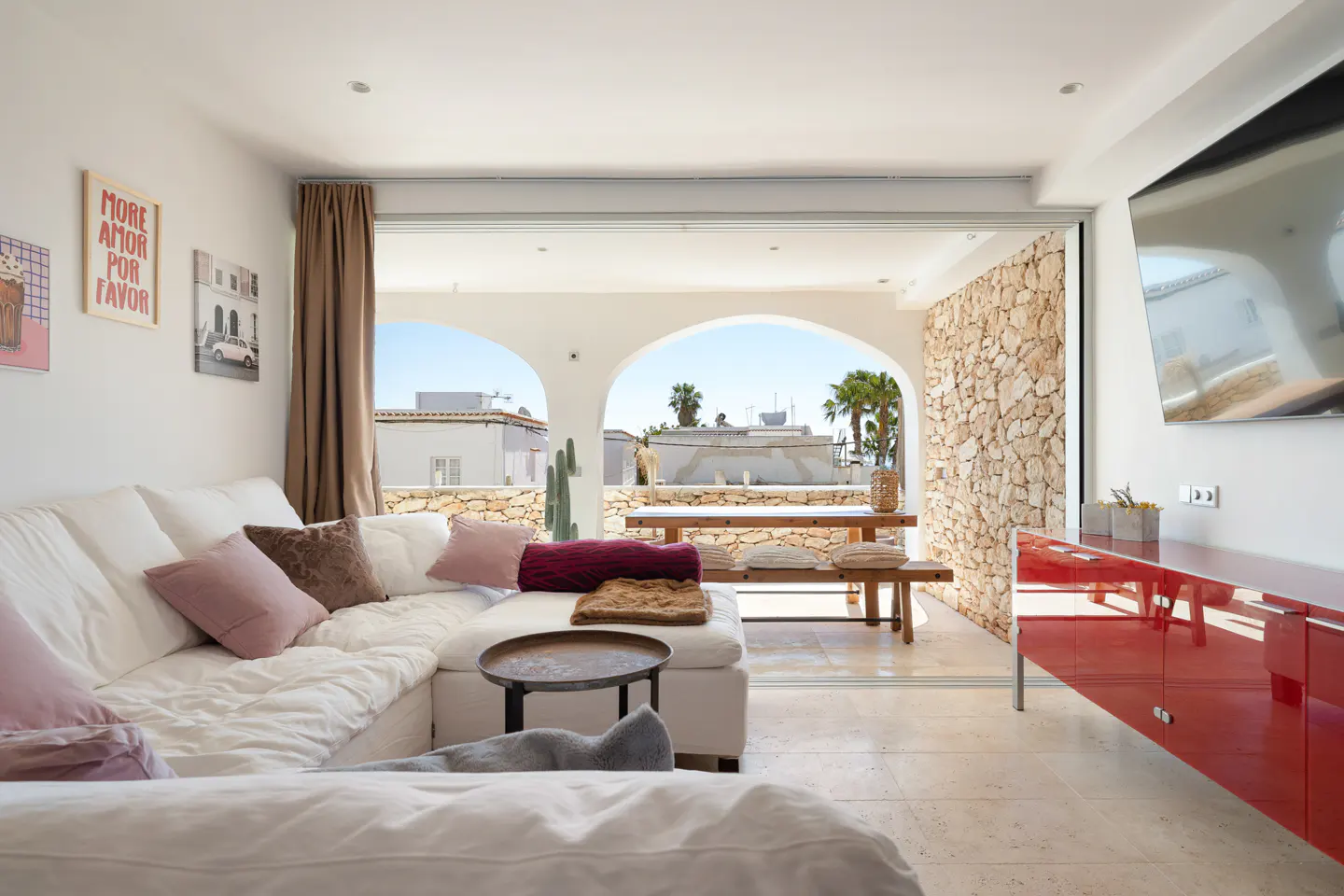 Bright living room with a white sofa, pink pillows, and a red cabinet. Arched windows lead to a stone patio with a wooden table.
