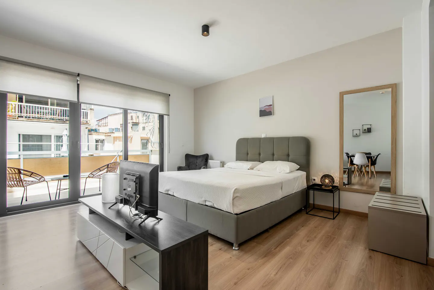 Bright bedroom with a gray bed, TV on a stand, and a balcony with chairs. A mirror reflects a dining area.