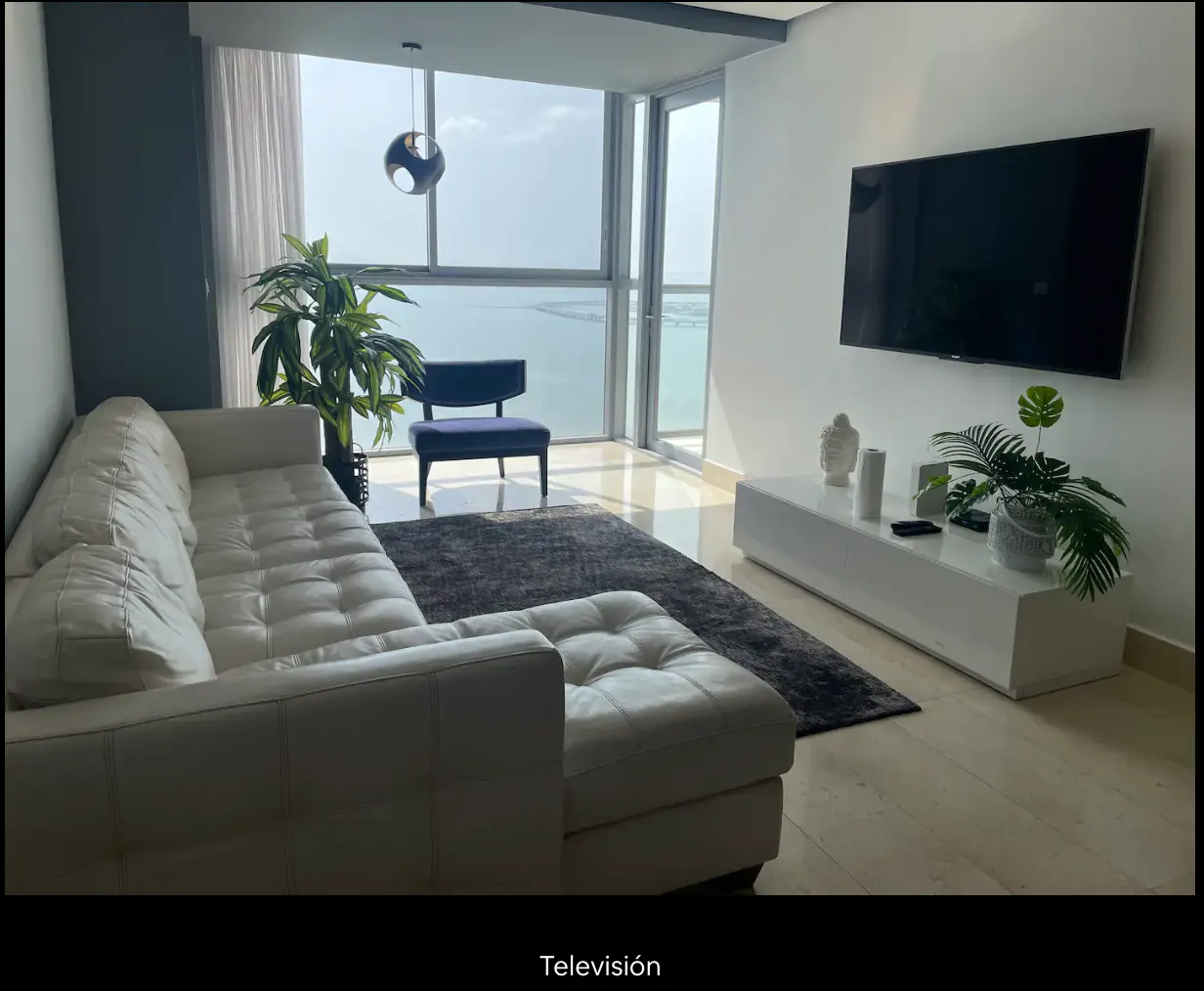 Living room with a white leather sofa, blue chair, and a large window with a water view. A TV hangs above a white console table.