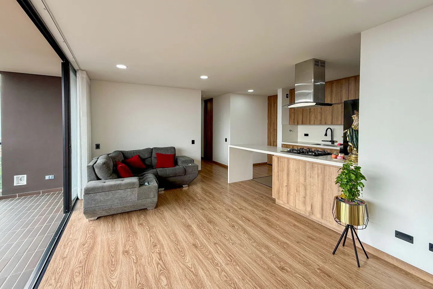 Modern open-plan apartment with a gray sectional sofa, wood-toned kitchen cabinets, and a balcony view.