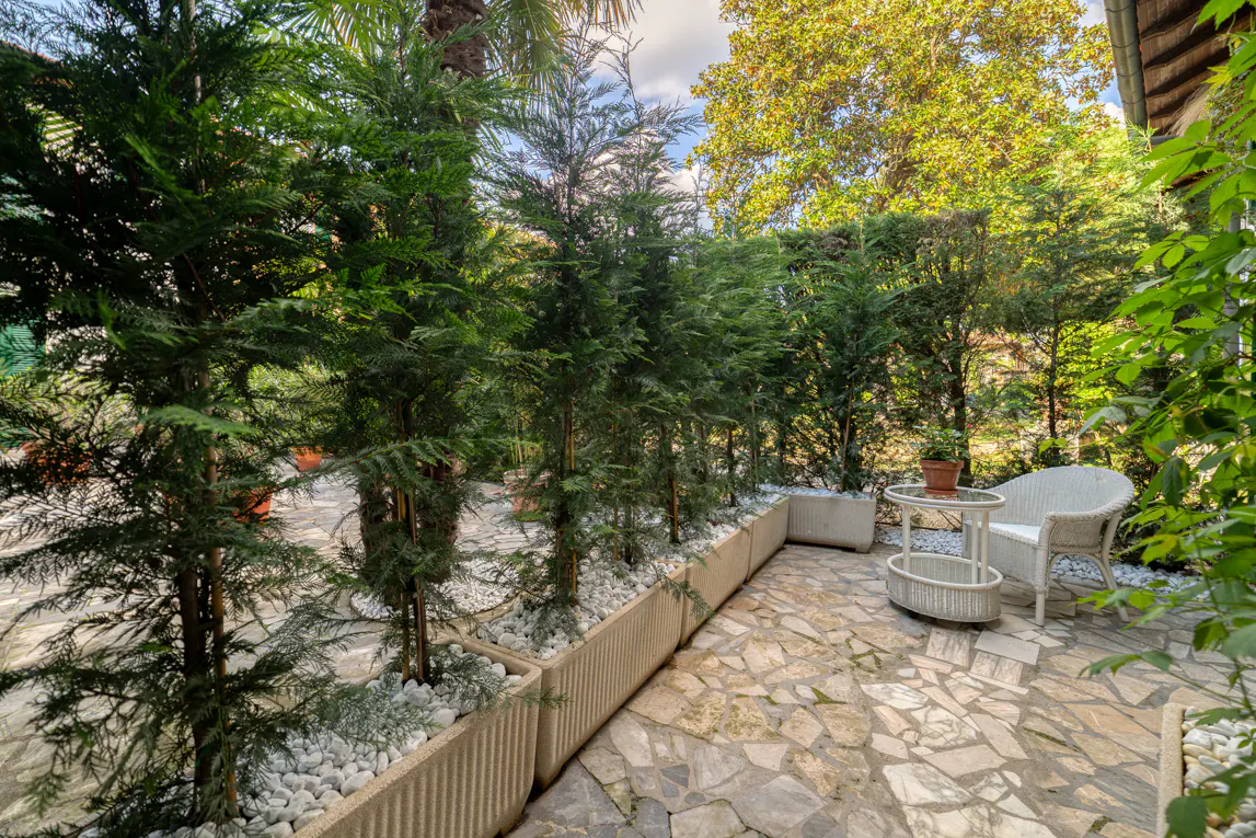 Outdoor patio with stone floor, white wicker chair and table, and tall green trees in planters.