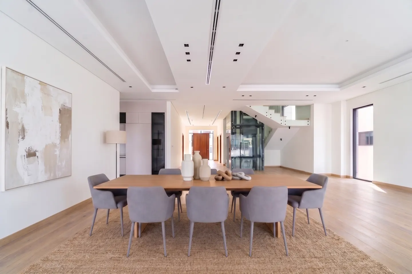 Bright dining room with a wood table, gray chairs, and a textured rug. Abstract art hangs on the white wall. A glass elevator and staircase are visible in the background.