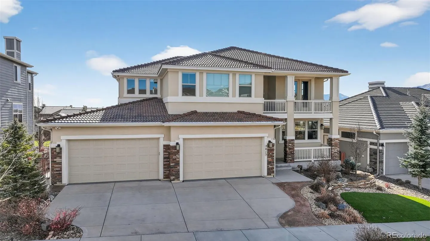 Two-story beige house with a brown tile roof, two-car garage, and a second-story balcony on a sunny day.