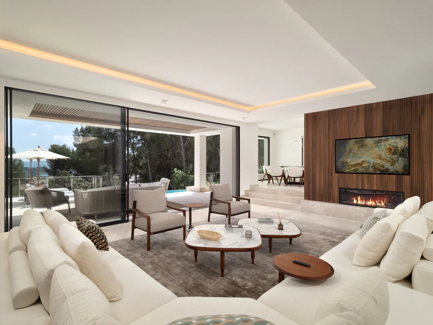 Bright living room with white sofa, chairs, and marble tables. A fireplace and TV are on a wood-paneled wall. Sliding doors open to a patio with ocean views.