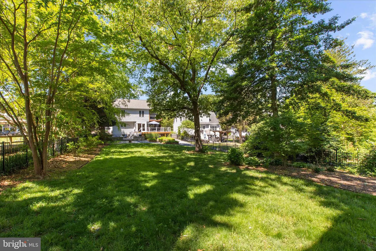 A large green lawn with trees casting shadows leads to a gray house with a deck and patio.