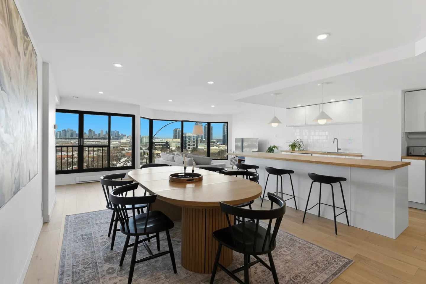 Bright, modern condo interior with a wood dining table, black chairs, and a white kitchen island. Large windows offer a city view.