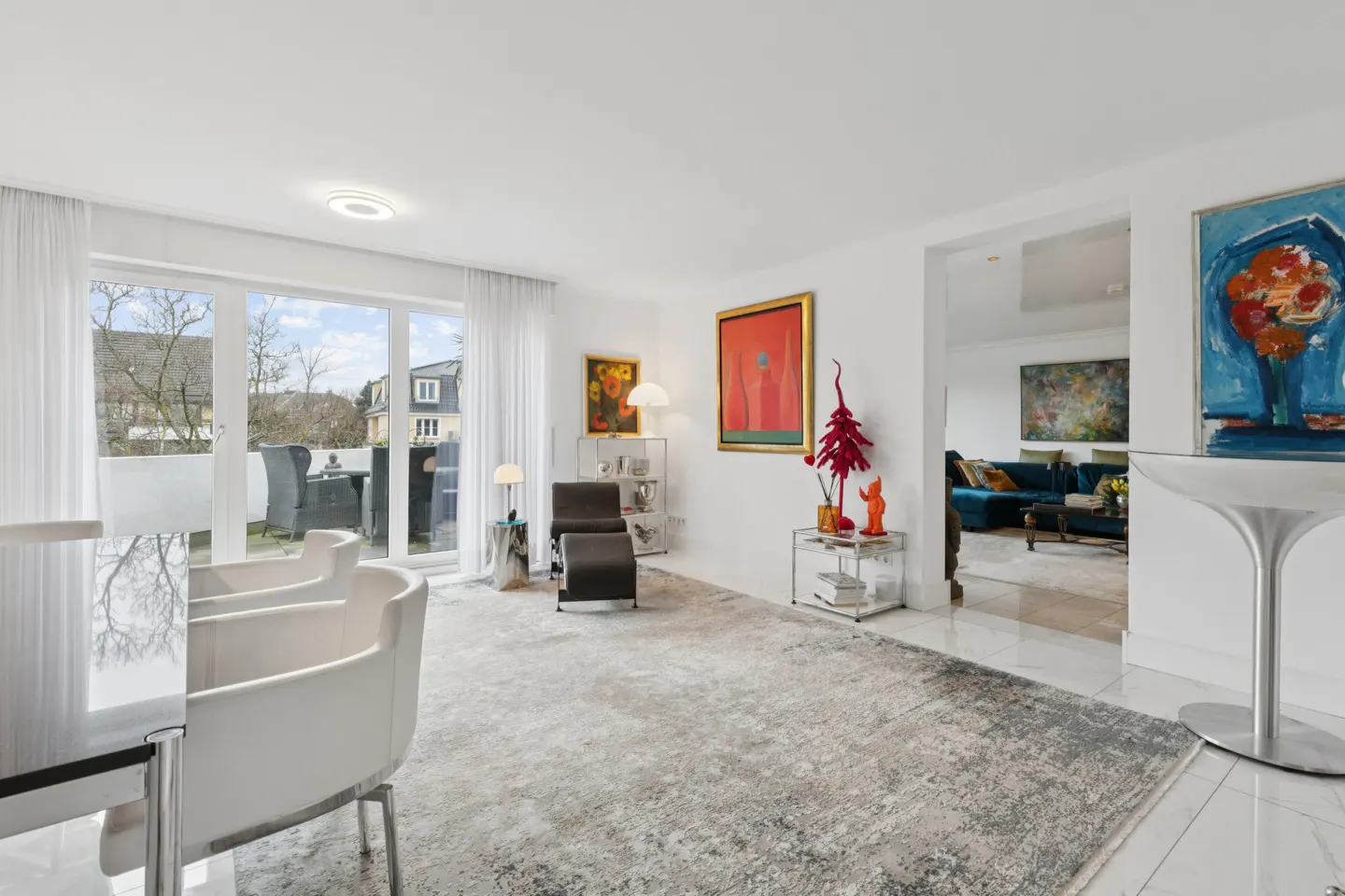 Bright, modern living room with white walls, a gray rug, and colorful art. A glass door leads to a balcony. A blue velvet sofa is visible in the next room.