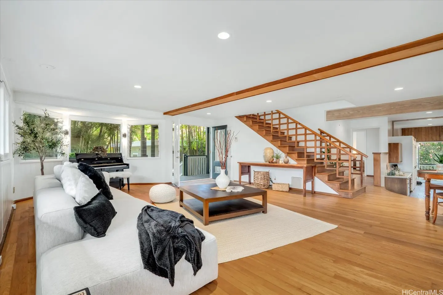 Bright living room with white sofa, black piano, and wooden staircase. Large windows offer a view of lush greenery.