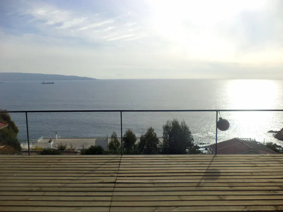 Ocean view from a wooden deck with a glass railing. Trees and houses are visible below. The sun reflects on the water.
