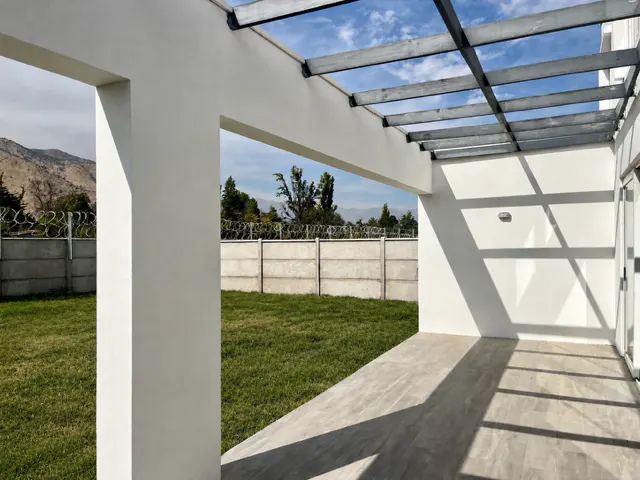 Modern patio with white walls, a gray pergola, and a concrete floor. Green lawn and a concrete fence with barbed wire are visible in the background.