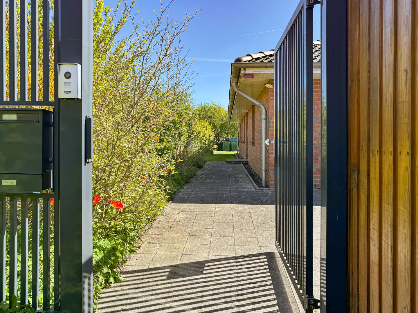 View through an open black metal gate to a brick house with a tiled roof and a paved walkway lined with greenery.