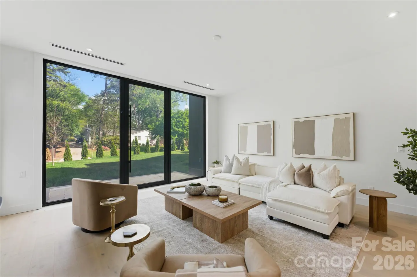 Modern living room with white sectional, tan armchairs, wooden coffee table, and floor-to-ceiling windows overlooking a yard.