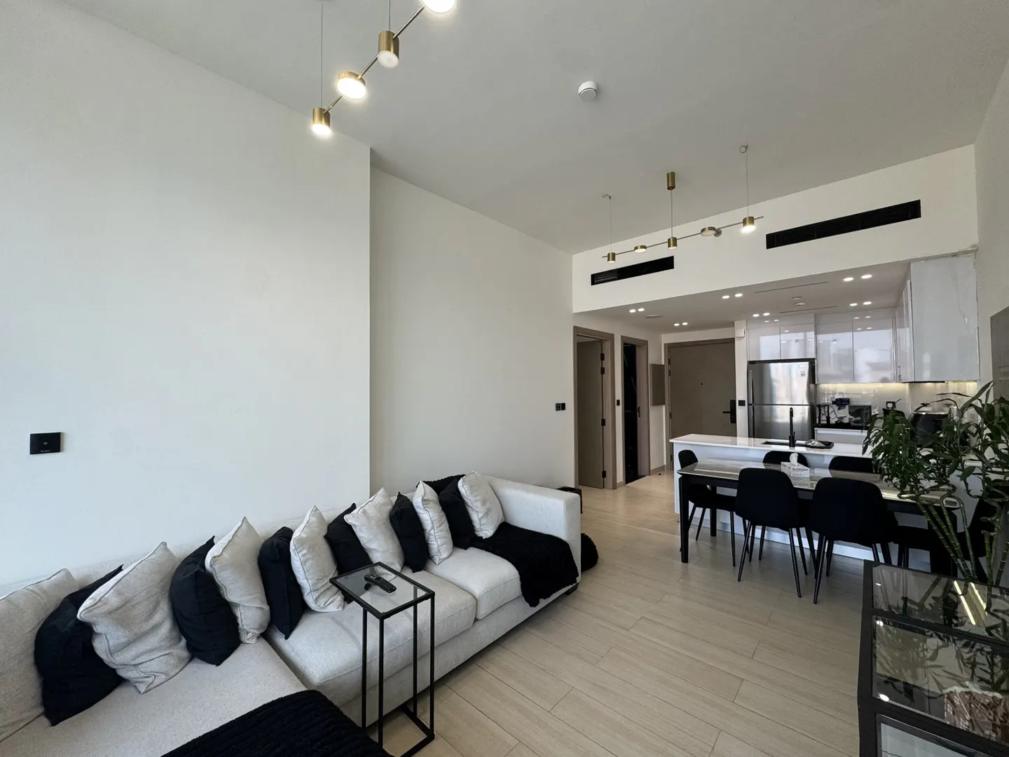 Bright, modern apartment interior with white walls, light wood floors, and a white sofa with black and gray pillows. A black dining table and chairs are visible in the kitchen area.