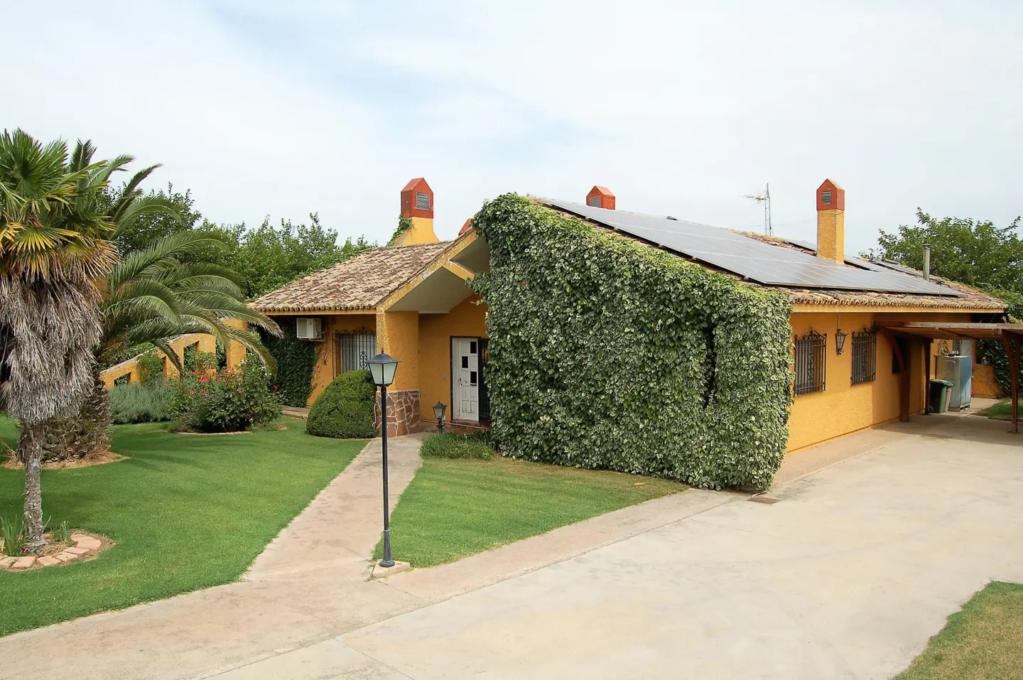 A yellow house with a tile roof and solar panels, partially covered in green ivy, sits on a green lawn.