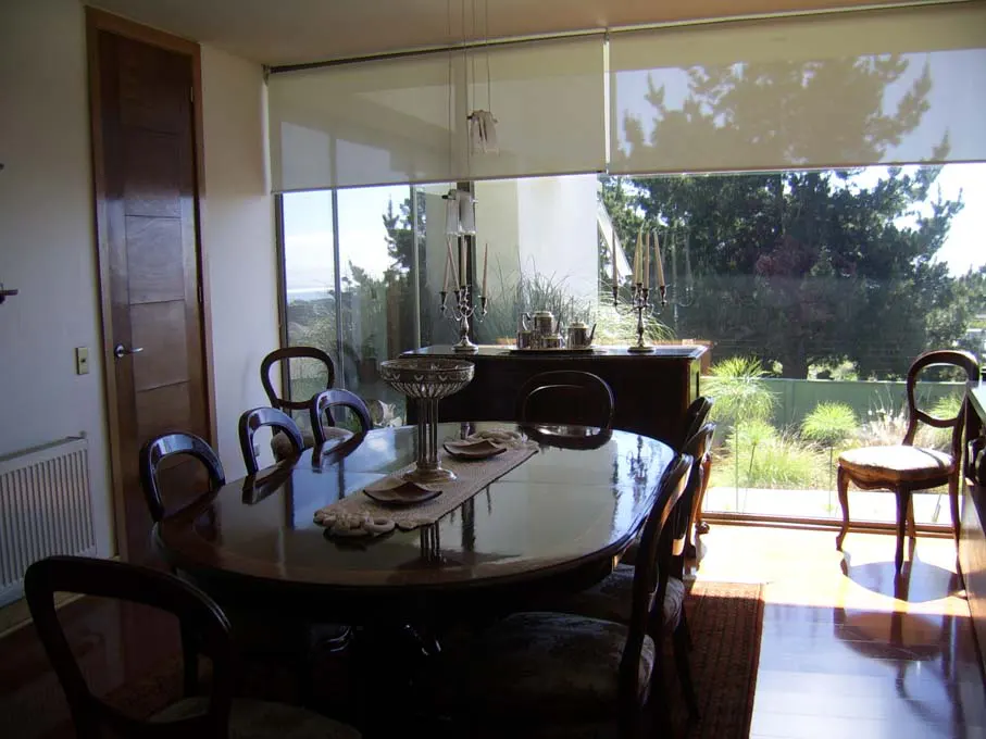 Dining room with dark wood table and chairs, large windows overlooking a green garden and ocean view.
