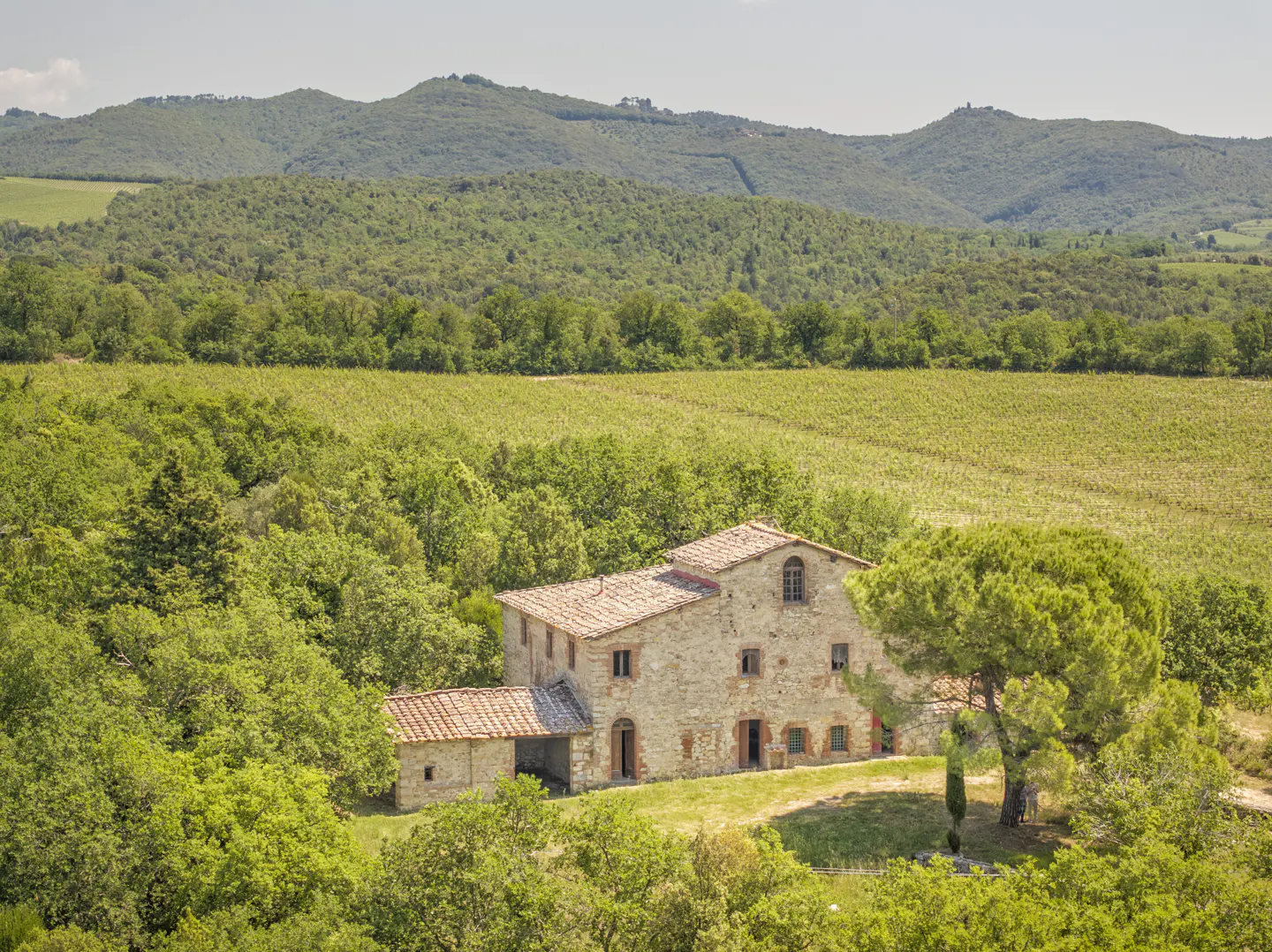 A rustic stone farmhouse with a terracotta roof nestled in a lush green landscape of trees, fields, and rolling hills.