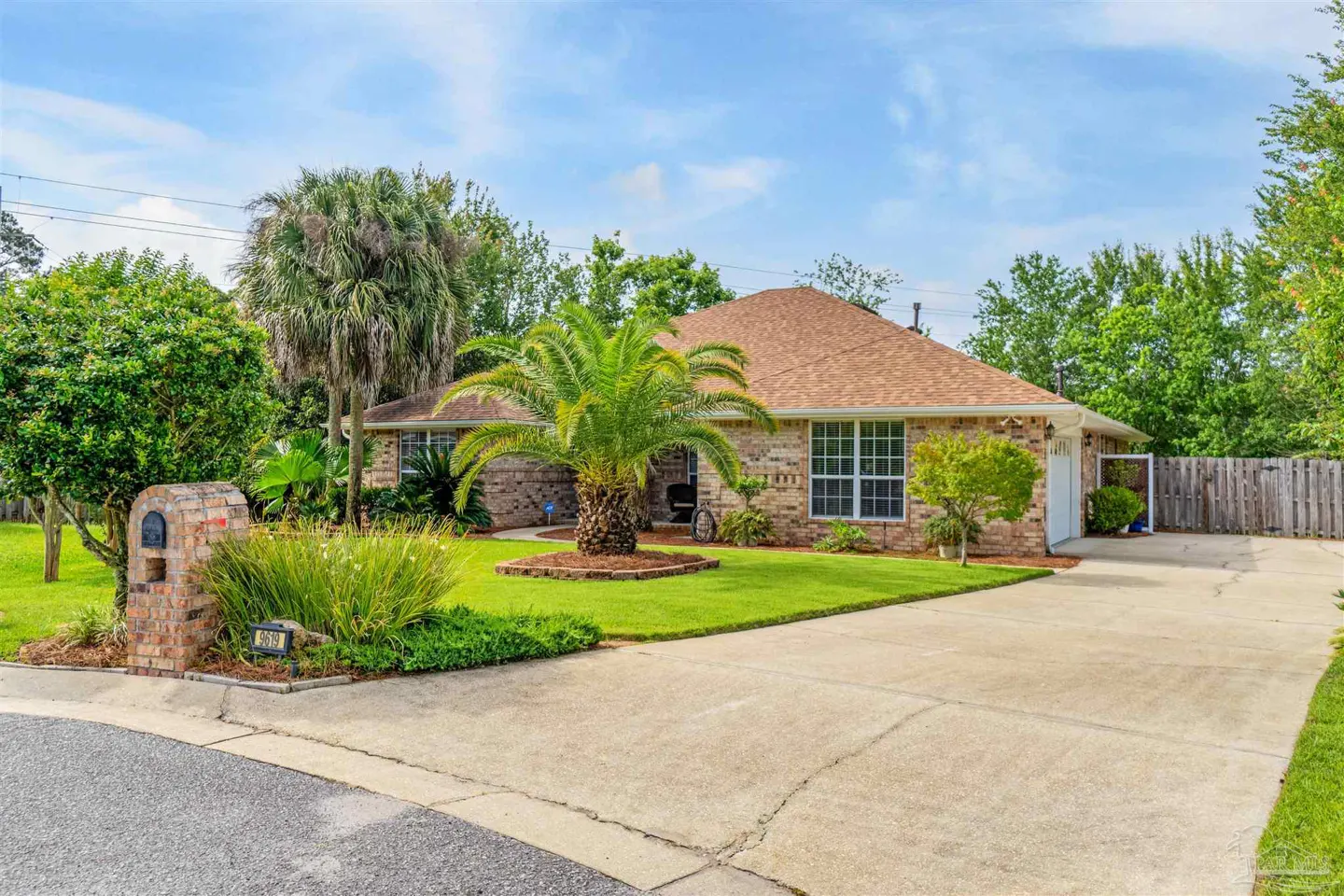 Single-story brick house with a tan shingle roof, palm trees, green lawn, and a concrete driveway under a blue sky.