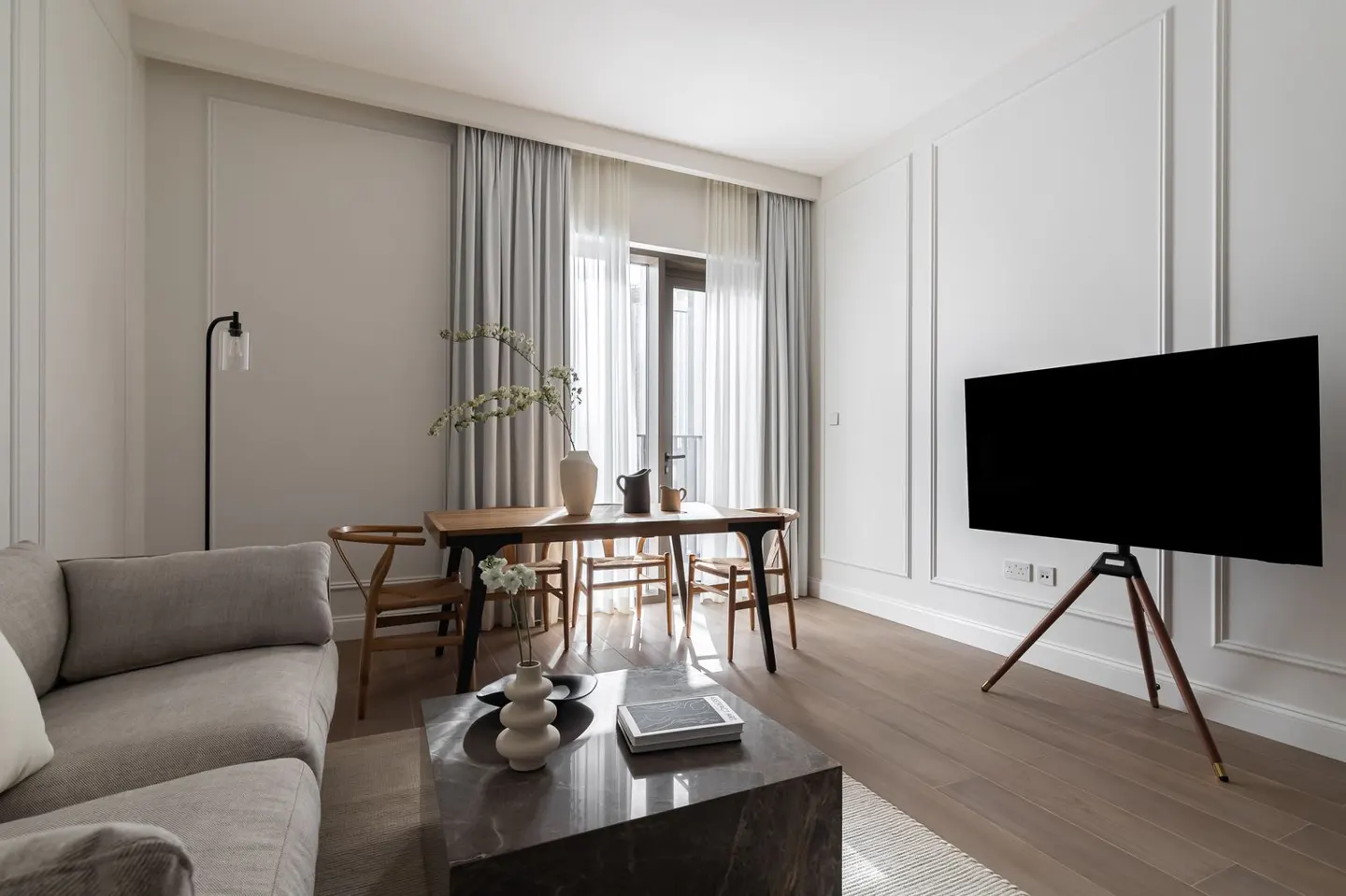 Living room with a gray sofa, marble coffee table, wood dining table with chairs, and a large black TV on a tripod stand.