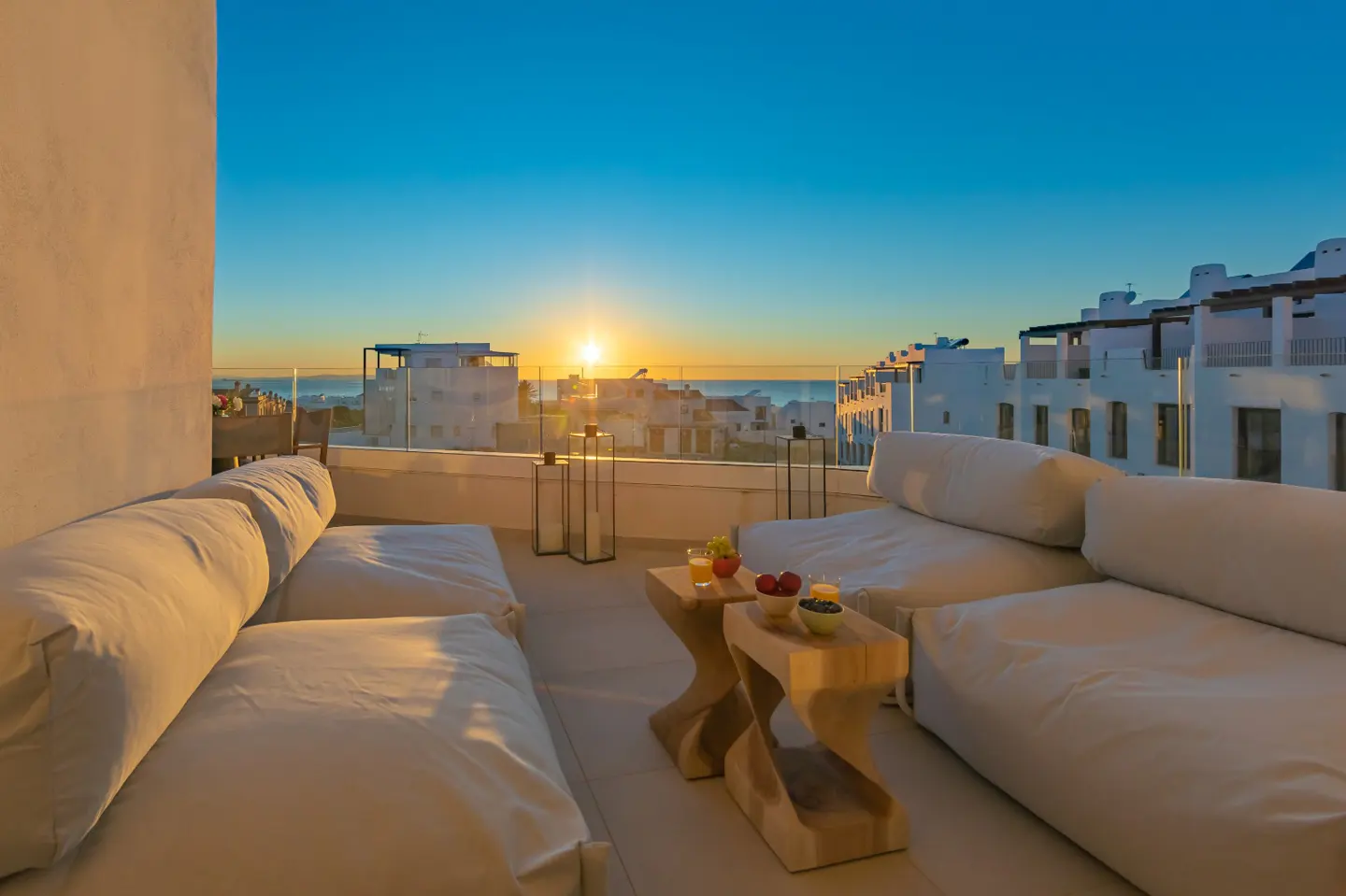 Balcony view with white sofas, wooden tables, and fruit. Ocean and sunrise in the background with white buildings.