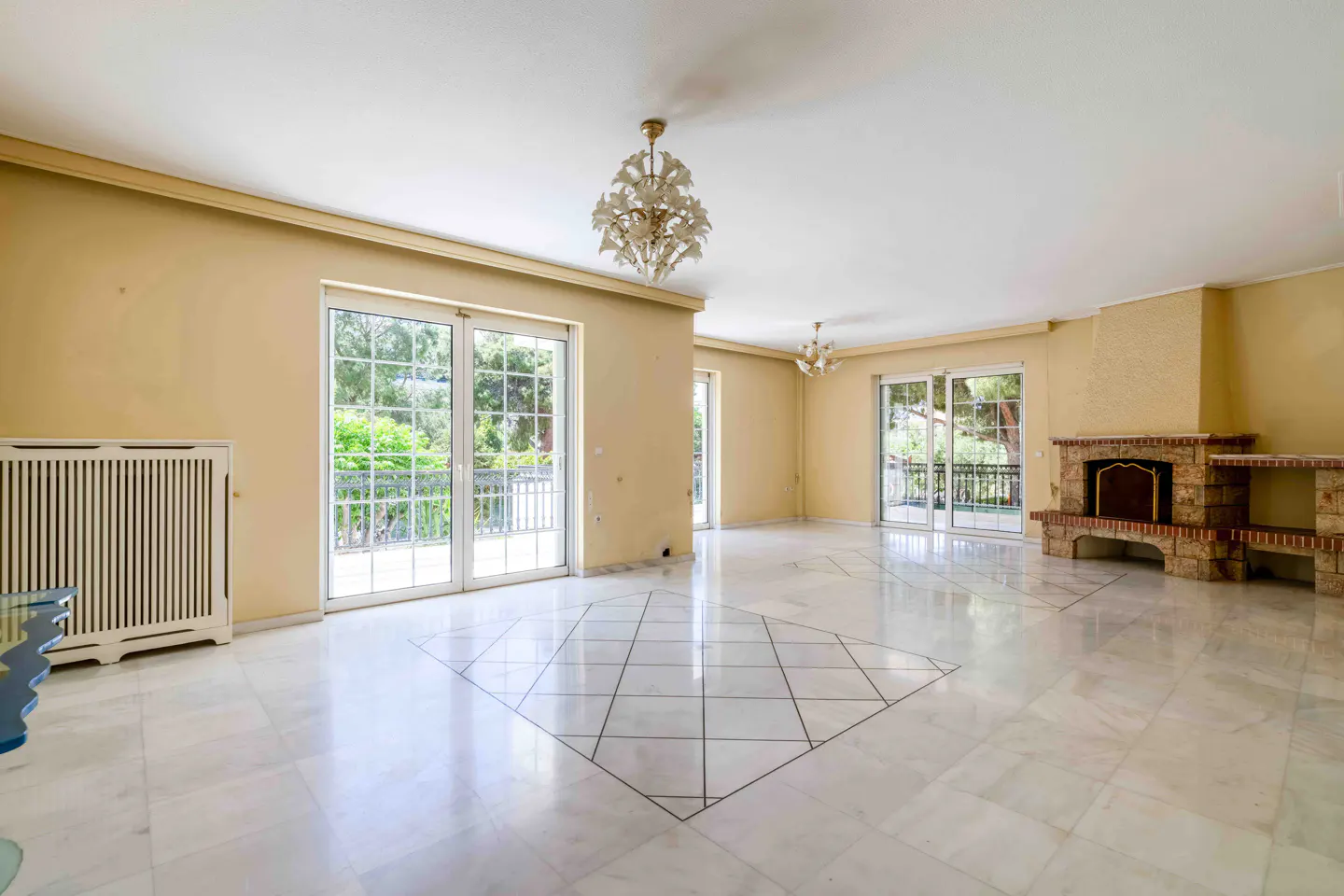 Spacious living room with marble floors, yellow walls, and a stone fireplace. Glass doors lead to a balcony with green trees visible.