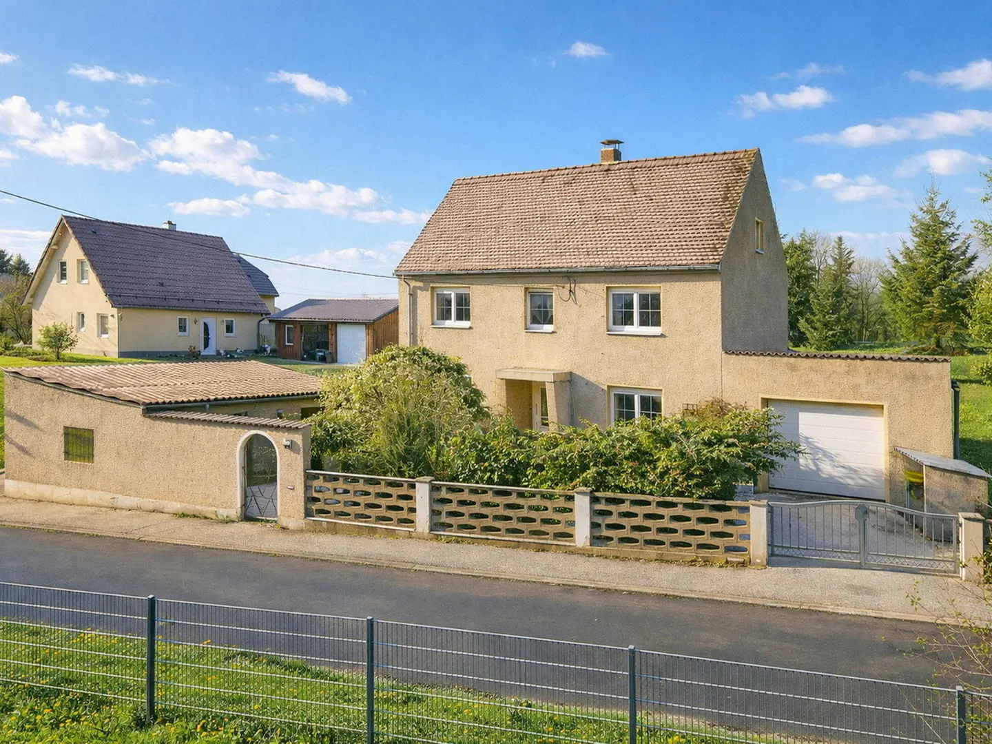 Two-story beige house with a brown roof, a garage, and a concrete fence on a sunny day.