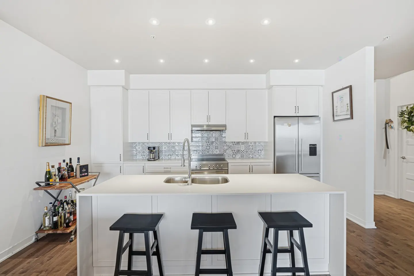 Bright, modern kitchen with white cabinets, stainless steel appliances, and a large island with three black stools. A bar cart sits to the left.