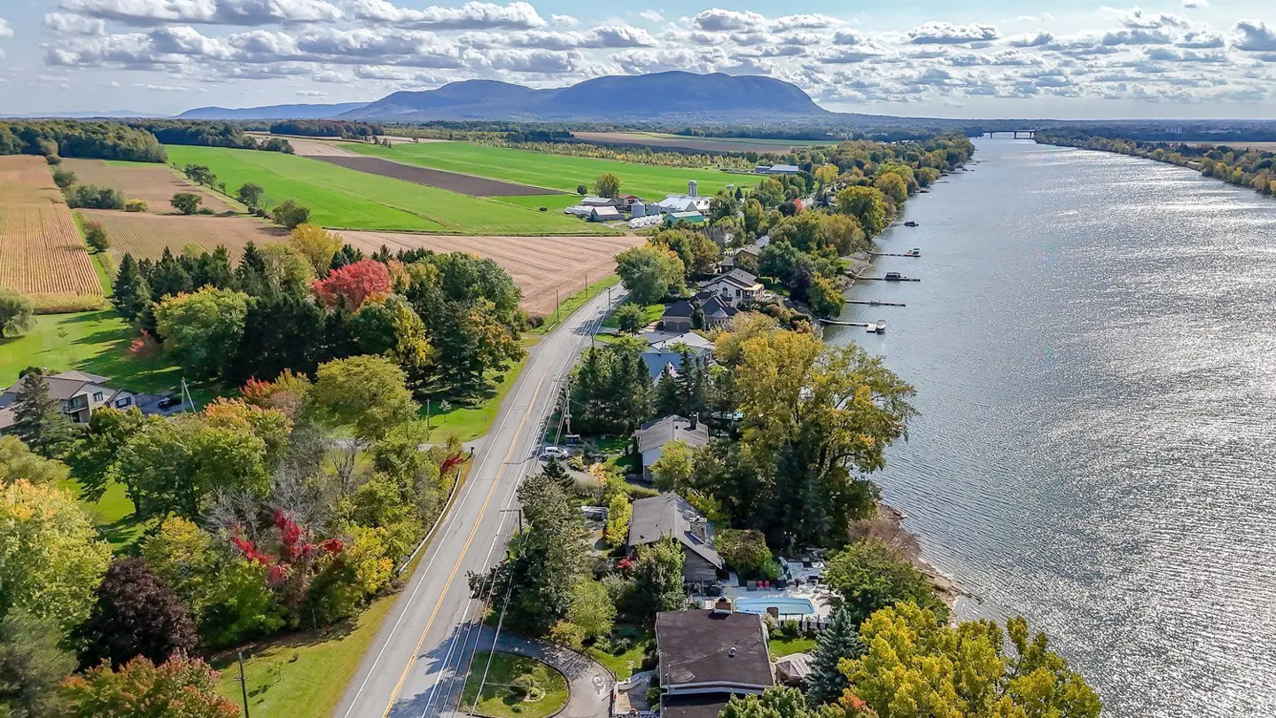 Aerial view of a river, fields, and homes on a sunny day. A road runs parallel to the river, with trees in fall colors. Mountains are visible in the distance.