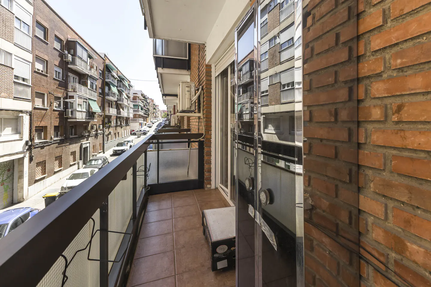 Narrow apartment balcony with tiled floor, brick wall, and glass railing overlooking a city street with parked cars.