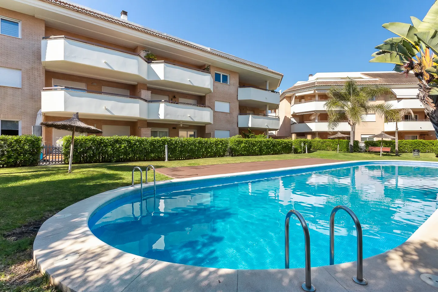 A turquoise pool with metal ladders sits in front of a brick apartment building with white balconies on a sunny day.