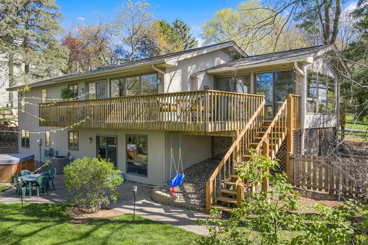 Beige two-story house with a wooden deck, stairs, and a blue swing. A patio with green furniture and a hot tub are visible.