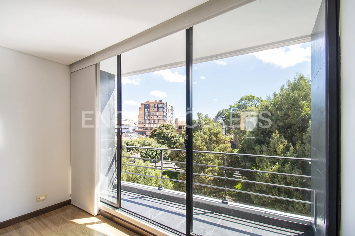 A bright room with a large sliding glass door leading to a balcony with a view of trees and a building.