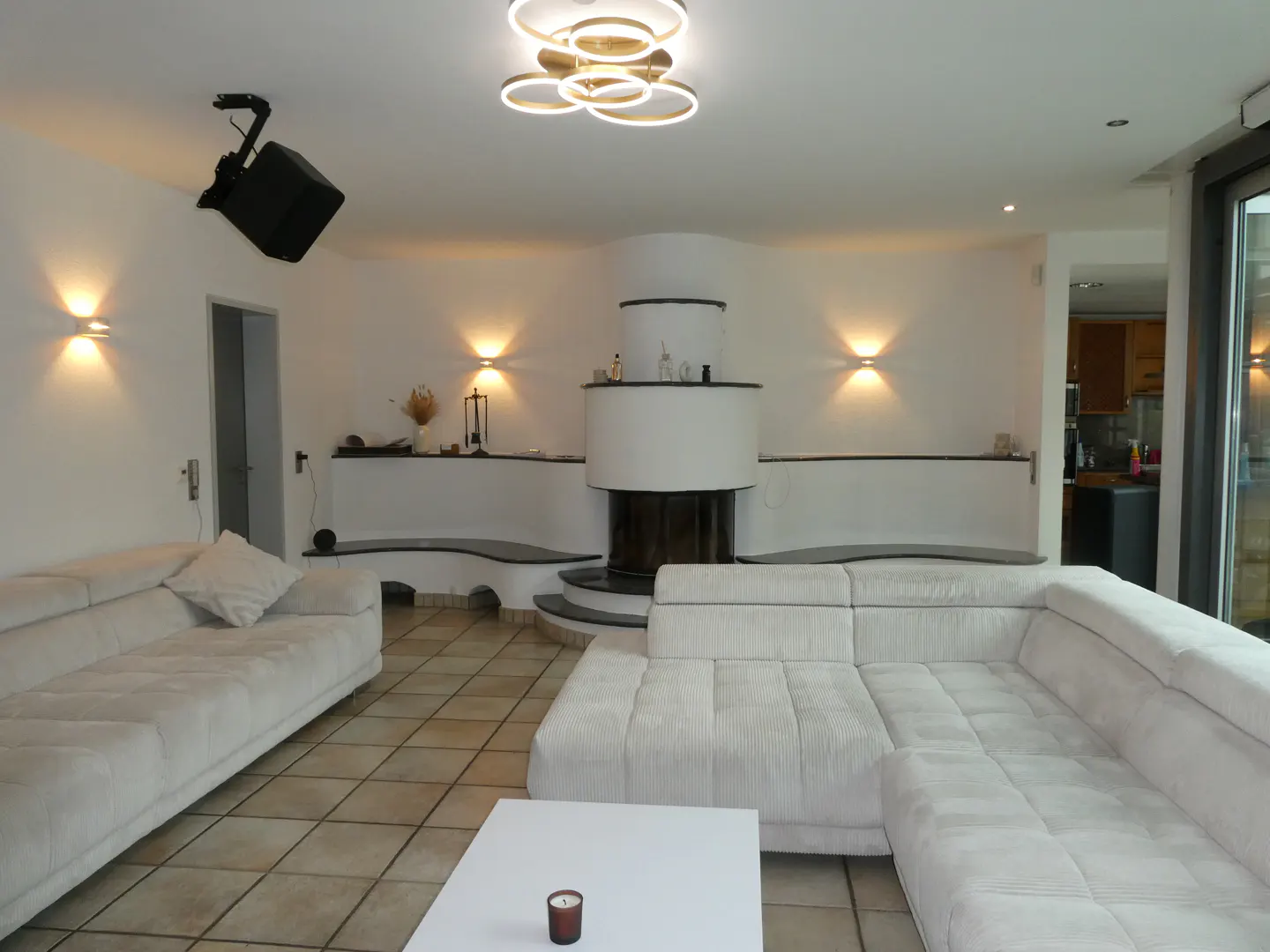 Living room with white sofas, a fireplace, and a modern chandelier. A black projector is mounted on the ceiling. Tiled floor.