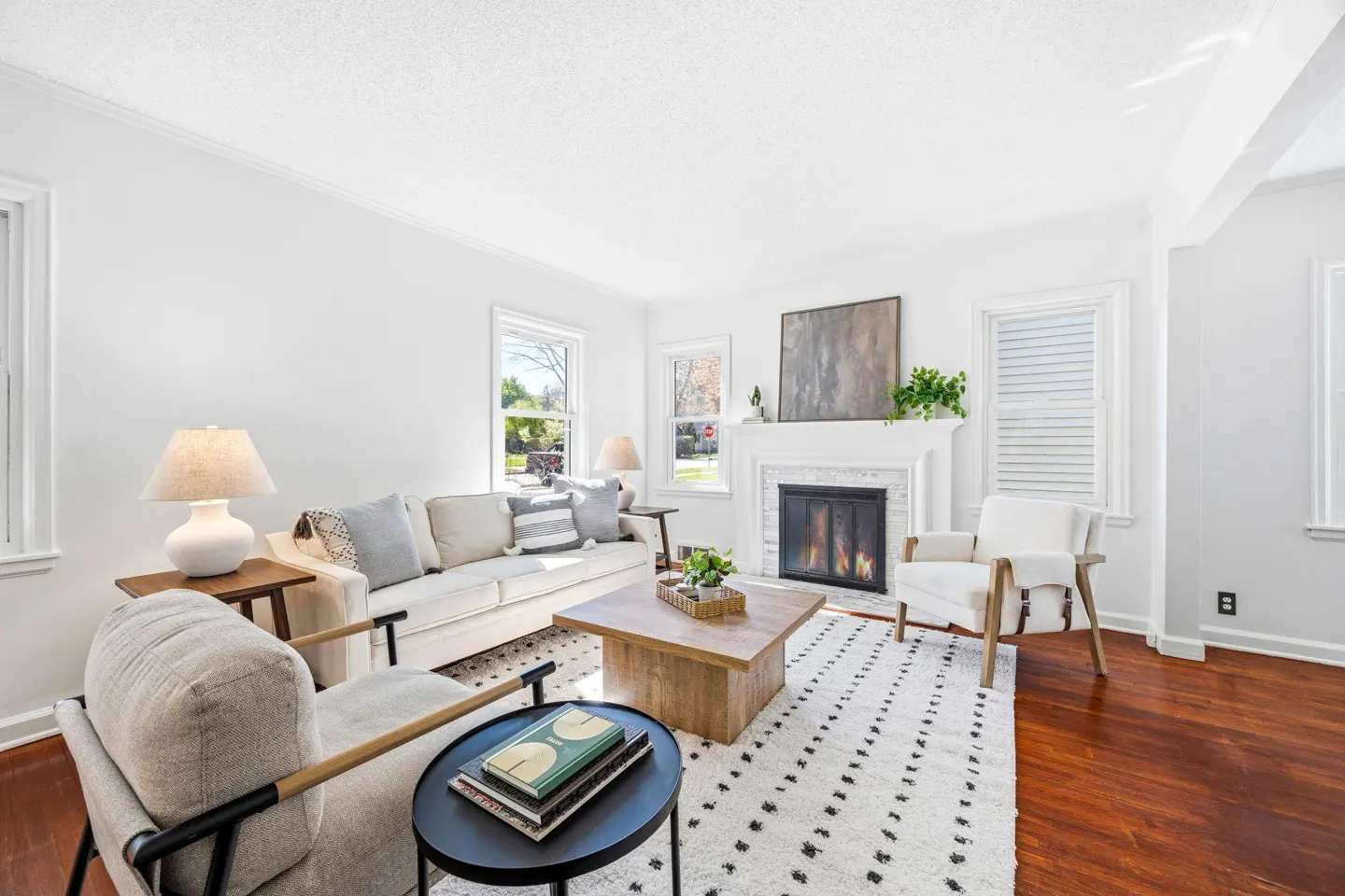 Bright living room with white walls, hardwood floors, and a fireplace. A white sofa and chairs sit on a patterned rug.