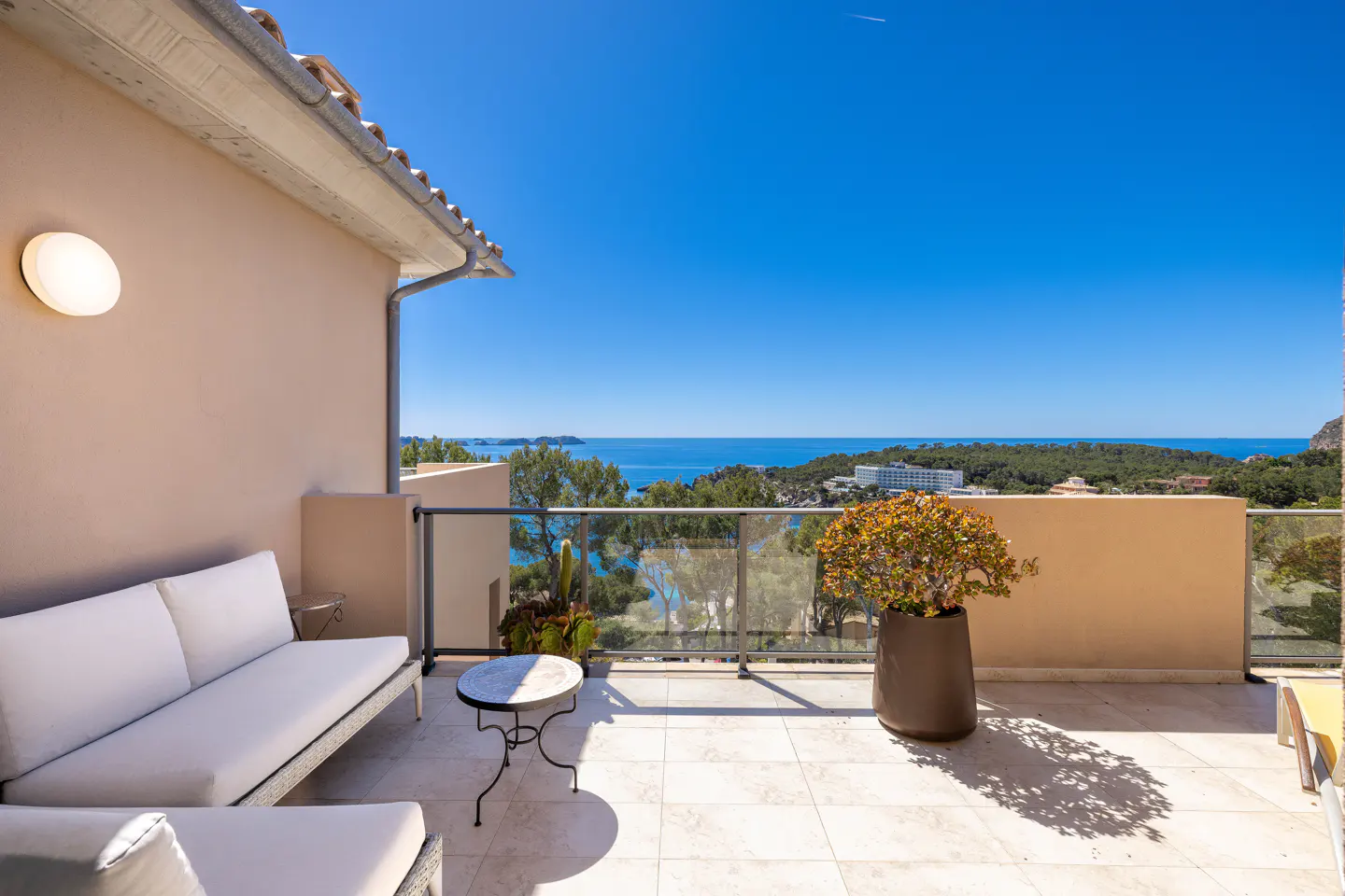 Balcony with white sofa, table, and potted plant overlooking the ocean on a sunny day.