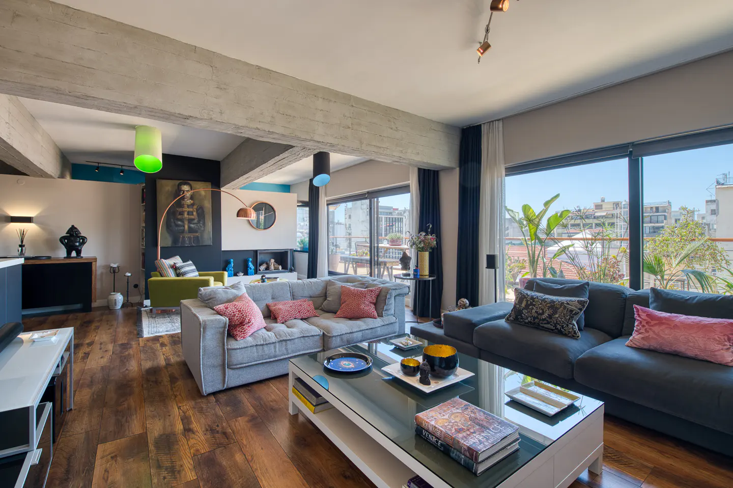 Bright living room with hardwood floors, gray sofas with pink pillows, and a glass coffee table. Large windows offer city views.
