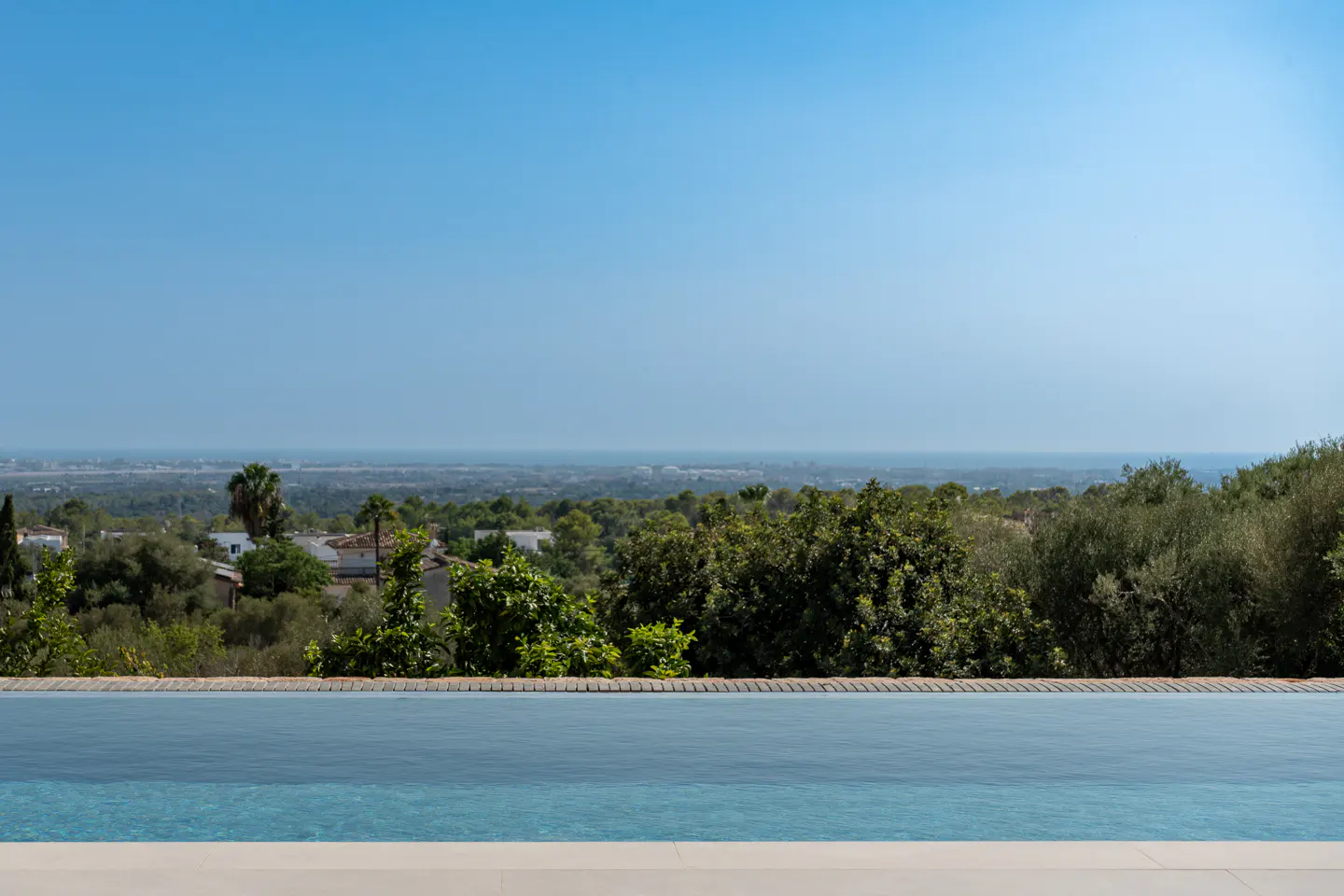 Infinity pool with blue water overlooking green trees, houses, and a distant city under a clear blue sky.